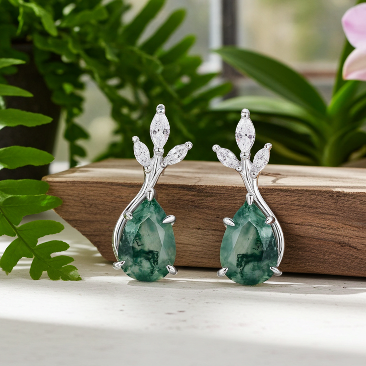 Pair of green teardrop earrings with silver accents on a wooden surface with plants in the background.